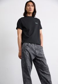 Camiseta de algodón negra con mangas enrolladas y logotipo turquesa, combinada con jeans grises que presentan un patrón intrincado y un ajuste relajado.