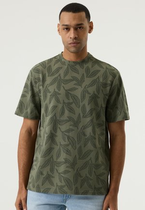 Mann mit grünem kurzärmligem Hemd mit Blattmuster und hellblauen Jeans, stehend vor einfachem hellem Hintergrund.