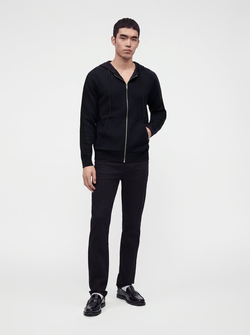 Young man standing wearing a black zip-up hoodie, black pants, and black dress shoes against a plain light background.