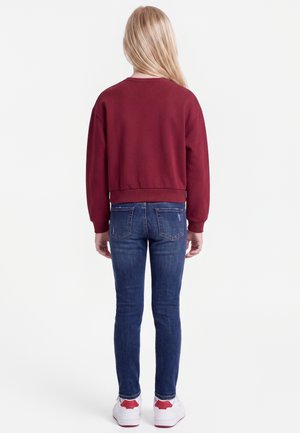 Enfant aux longs cheveux blonds, vu de dos, portant un sweat-shirt bordeaux, un jean bleu et des baskets blanches avec des touches rouges.