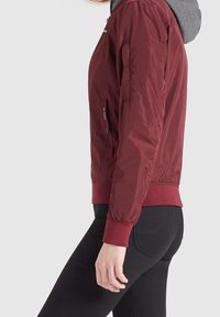 Vínově červená bomber bunda se šedou kapucí, elastickými manžetami a kapsou na zip. Hladká textura a přiléhavý střih.