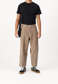 Beige kordflöjlsbyxor med hög midja, lös passform, i kombination med en svart t-shirt med kort ärm och svart-vita sneakers. Justerbar axelväska.