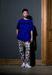 T-shirt azul oversize com o texto "Moschino Couture", combinado com calças de padrão gráfico colorido e ténis brancos. Textura suave, ajuste casual.