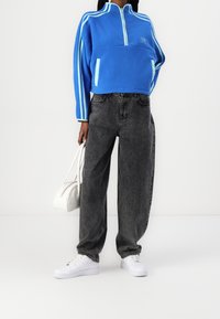 Pullover in pile blu con zip a metà e tasche laterali, caratterizzato da strisce azzurre. Abbinato a jeans neri a vita alta e a gamba larga e sneakers bianche.