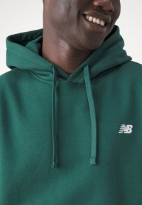 Žalias medvilninis gobtuvėlis su kengūros kišene, reguliuojamais raiščiais ir baltu išsiuvinėtu „New Balance“ logotipu krūtinėje.