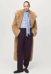 Femme portant un long manteau en fausse fourrure beige, une blouse bleu clair avec un col noué, un pantalon large violet foncé et des mocassins marron avec des chaussettes blanches.