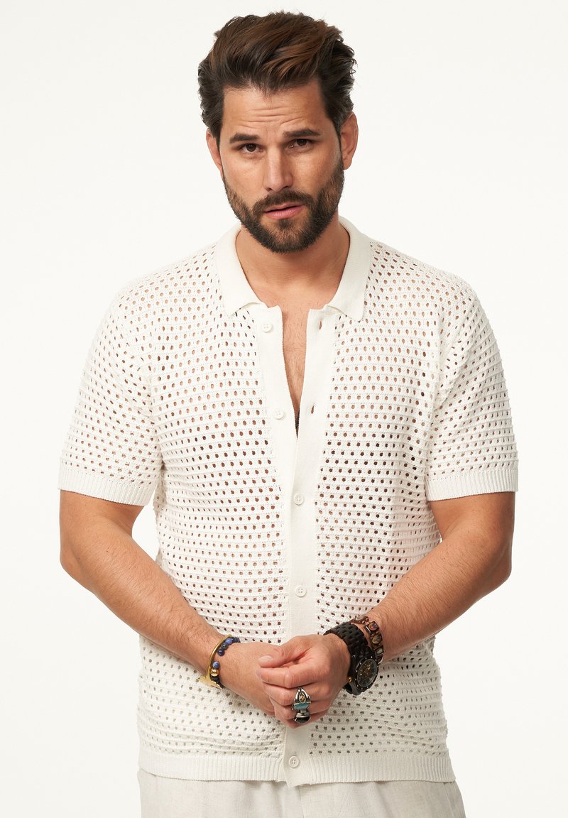 Kurzärmeliger, knöpfbarer Cardigan in hellem Creme, mit einem Spitzenstrickmuster, gerippten Bündchen und einem Kragen. Braune Armbänder an den Handgelenken.