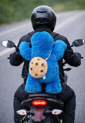 Motociclista vestito con abbigliamento nero guida su strada con uno zaino grande di peluche blu a forma di Cookie Monster con motivo biscotto.