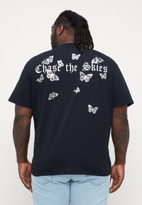 Svart bomull T-skjorte med hvite sommerfugltrykk spredt over ryggen, med teksten "Chase the Skies" over designet. Korte ermer.