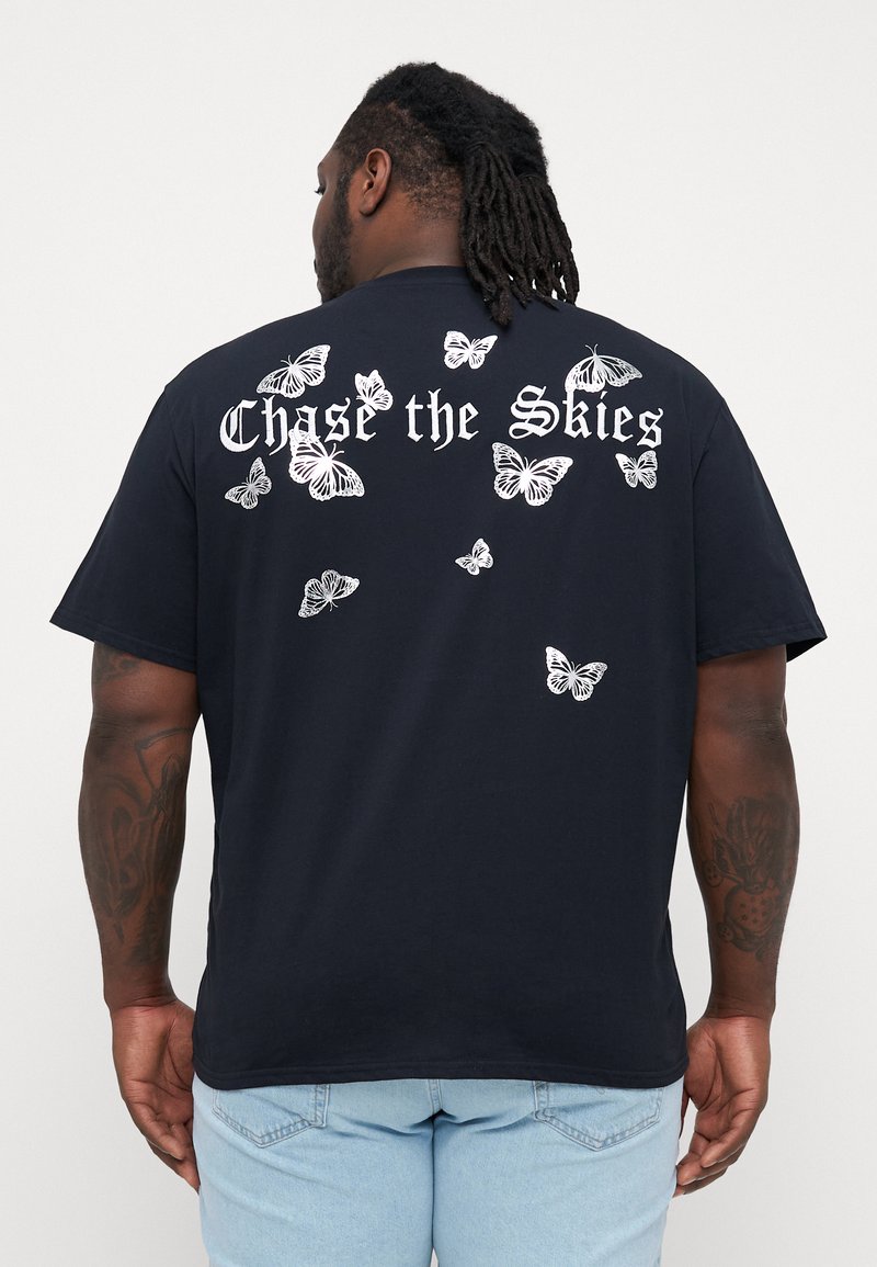 Svart bomull T-skjorte med hvite sommerfugltrykk spredt over ryggen, med teksten "Chase the Skies" over designet. Korte ermer.