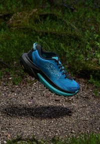 Zapatilla de trail azul con una parte superior texturizada, suela negra y acentos turquesa. Diseño elevado con cordones visibles y logo de la marca en el lateral.