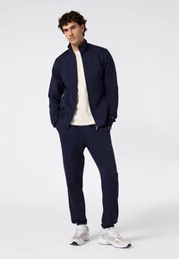 Marineblauw trainingspak van gladde stof, met een jack met rits en een broek met elastische tailleband. Accentlogo op het jack. Witte sneakers.