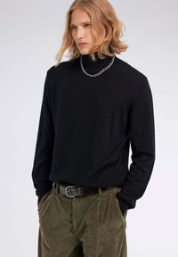 Schwarzer langärmeliger Rollkragenpullover mit gerippter Textur, kombiniert mit olivgrünen Cordhosen und einer klobigen silbernen Kettenhalskette.