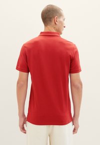 Rød polo t-shirt med korte ærmer, subtil tekstur, kravedetalje og sideskår i kanten, set fra ryggen mod en lys baggrund.