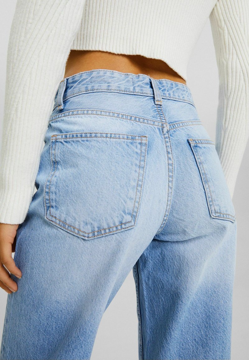 Un jean en denim bleu clair avec taille haute, coupe droite et deux poches arrière avec des coutures visibles. Tissu texturé avec un léger effet délavé.
