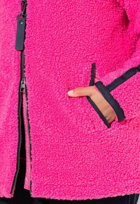 Vaaleanpunainen fleece-takki, jossa on teksturoitu pinta, vetoketjukiinnitys edessä, sivutaskut ja mustat yksityiskohdat hihansuissa ja taskuissa.