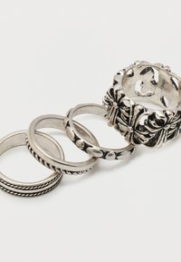 Σετ από τέσσερα ασημένια δαχτυλίδια με διαφορετικές υφές και σχέδια, συμπεριλαμβανομένου ενός φαρδιού δαχτυλιδιού με λουλουδένια μοτίβα, τοποθετημένα σε σειρά σε λευκό φόντο.