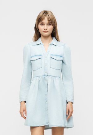 Ung kvinde iført en lyseblå langærmet denimkjole med knapper foran og to brystlommer, stående mod en ensfarvet baggrund.