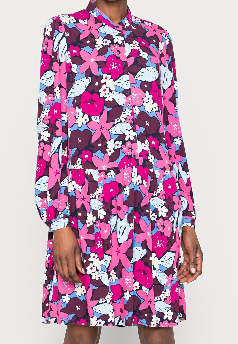 Robe à manches longues avec boutons et motif floral lumineux en rose, violet, bleu et blanc, portée par une personne à la peau foncée.