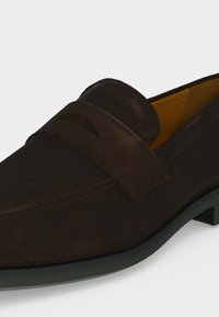 Σκούρος καφέ ανδρικός μοκασίνι από σουέντ με λουράκι πάνω από το επάνω μέρος, χαρακτηριστικό στρογγυλεμένη μύτη και μαύρη σόλα.