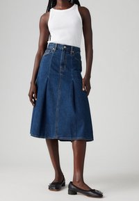 Falda de mezclilla en azul medio con frente plisado, cintura alta y corte hasta la rodilla; combinada con una camiseta sin mangas blanca y zapatos planos negros.