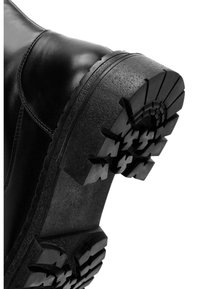 Stivale in pelle nera con suola in gomma resistente, caratterizzata da scanalature profonde e un motivo testurizzato. Il design è angolare e robusto.
