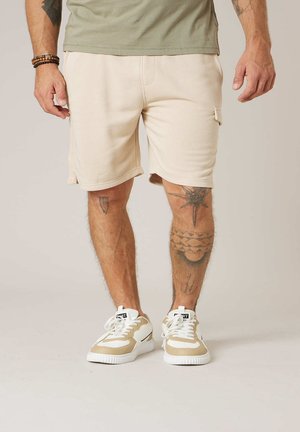 Homme portant un short cargo beige, des baskets blanches et beiges, ainsi que des bracelets, debout avec des tatouages visibles sur les deux jambes sur un fond neutre.