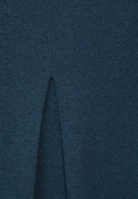 Stickad mörk teal tyg med en subtil textur har en delad detalj längst ner, vilket skapar ett distinkt formande inslag.