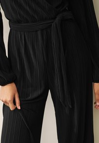 Jumpsuit noir côtelé avec des manches longues et une ceinture à nouer. Le tissu a un motif de rayures verticales texturé. Gros plan sur la taille et la manche.