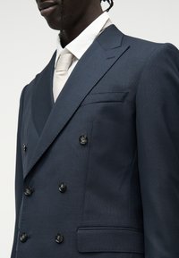 Homme noir portant une veste de costume croisée bleu marine foncé avec des boutons noirs, une chemise blanche et une cravate beige clair.