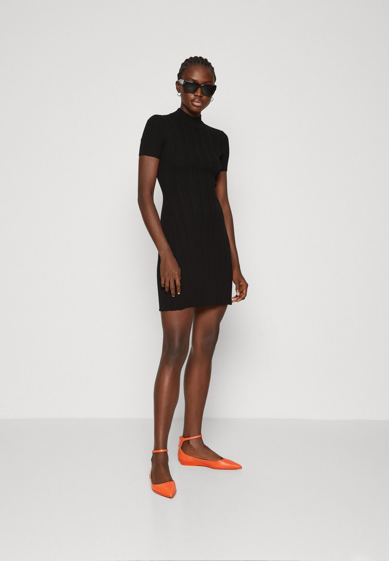 Schwarzes geripptes Strickkleid mit kurzen Ärmeln und hohem Kragen, kombiniert mit leuchtend orangefarbenen flachen Schuhen mit Knöchelriemen.