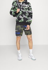 Felpa nera con grafiche verdi e bianche, abbinata a pantaloni corti neri con motivi geometrici blu e verdi, e sneakers bianche.