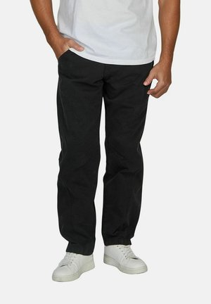 Man wearing black pants and white sneakers, with one hand in pocket and white t-shirt visible from waist down.