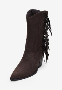 Botín de ante marrón con detalle de flecos, punta afilada y tacón apilado. Cierre con cremallera en el lado interior para fácil colocación.