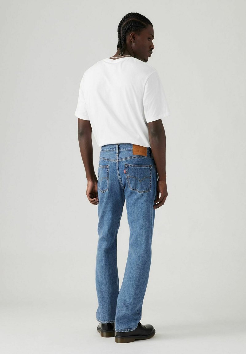 Vaqueros azules de mezclilla con piernas ligeramente ajustadas, un parche de cuero marrón en la parte trasera, combinados con una camiseta blanca lisa y zapatos negros.