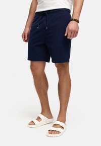 Bermudas texturizadas azul marino con cintura elástica y cordón. Combinadas con sandalias blancas que tienen una sola correa con hebilla.