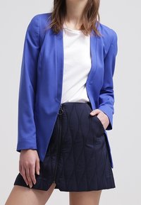Blazer azul con solapas con muesca, llevado sobre una blusa blanca y una falda oscura con un patrón de olas y un zipper en el frente.