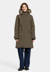 Brun parka med kunstig pelskant på hætten, lange ærmer, sidelommer og trykknaplukning. Løs pasform, strækker sig til midt på låret, parret med vide jeans.