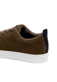 Zapatilla de ante marrón con suela de goma blanca, presenta una pestaña en el talón de color azul oscuro y cordones a juego, textura suave y diseño minimalista.