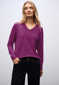 Pull-over violet à col en V fabriqué en tissu doux et texturé, associé à un jean foncé. Le pull a des manches longues et des poignets côtelés.