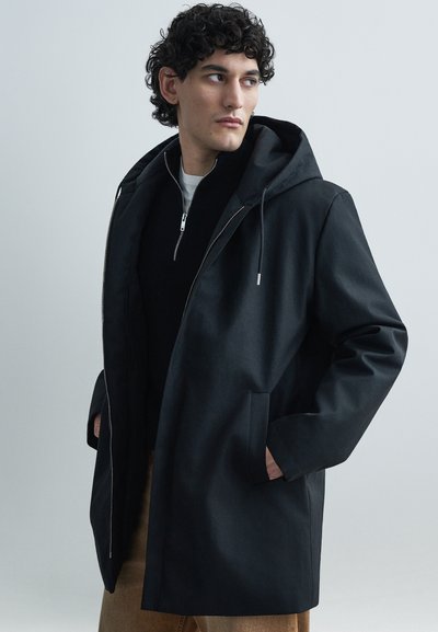 Veste noire à capuche avec une texture lisse, dotée d'une fermeture éclair, de poches latérales et d'une coupe décontractée. Portée sur un haut foncé.