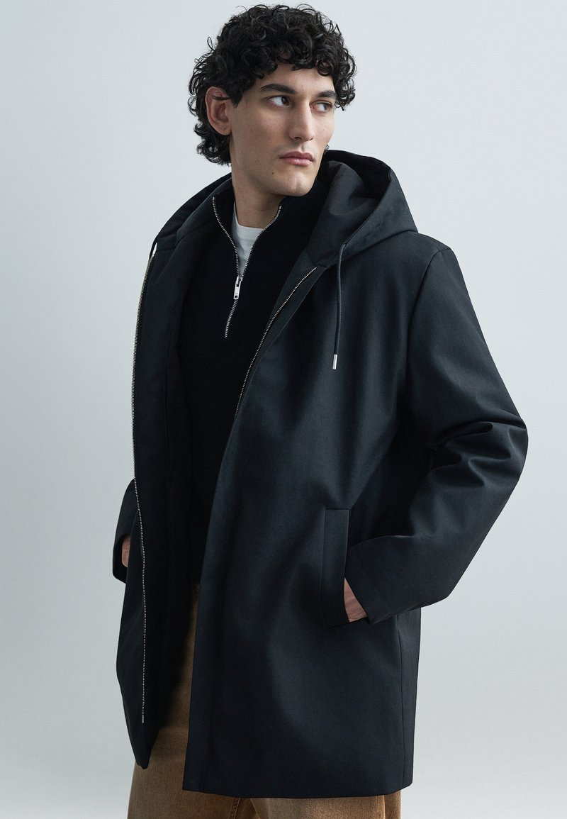 Must kapuutsiga jakk, millel on sile tekstuur, esikinnitus tõmblukuga, küljetaskud ja lõdvestunud istuvus. Kandatud tumeda topi peal.