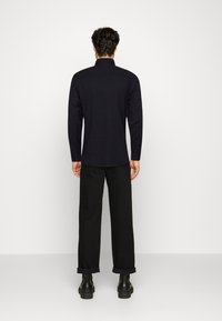 Černá košile s dlouhým rukávem a límečkem, navržená ve vypasovaném stylu. Kombinována s volnými černými kalhotami a kotníkovými botami. Jednoduchý, elegantní design.