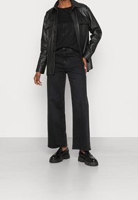 Svart läderjacka med två bröstfickor, kombinerad med vida svarta jeans och kraftiga svarta skor. Slät textur och minimal design.