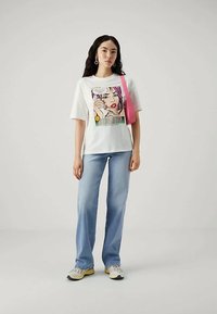 Νεαρή γυναίκα με μακριά σκούρα μαλλιά φοράει λευκό γραφικό T-shirt, ανοιχτό μπλε τζιν, λευκά αθλητικά παπούτσια, σκουλαρίκια σε σχήμα καρδιάς και ροζ τσάντα ώμου.