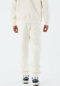 Mann in cremefarbenen Jogginghose und Sweatshirt mit schwarz-weißen Turnschuhen, stehend mit dem Rücken zu einem weißen Hintergrund.