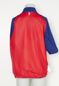 Americana deportiva con espalda de malla roja, mangas azules y cuello. Presenta un dobladillo curvado y aberturas laterales para ventilación. Etiqueta visible en el lateral.