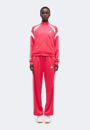 Person steht in einem rot-weißen Adidas Trainingsanzug mit weißen Sneakers, schaut nach vorne vor einem schlichten hellen Hintergrund.