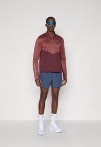 Hombre vestido con un pullover de cremallera corta burdeos, pantalones cortos Nike azules, calcetines blancos y zapatillas deportivas, y gafas de sol deportivas reflectantes azules de pie contra un fondo blanco.