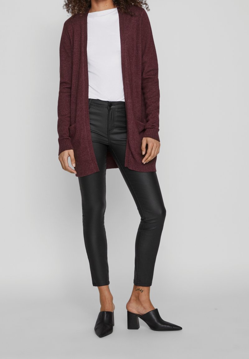 Femme portant un cardigan bordeaux sur un top blanc, un pantalon skinny en similicuir noir et des mules noires à talons, debout devant un fond gris.